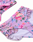 A close-up of a children's bikini bottom in light lavender with a colorful floral print, featuring ruffled trim along the leg openings, shown on a white background.