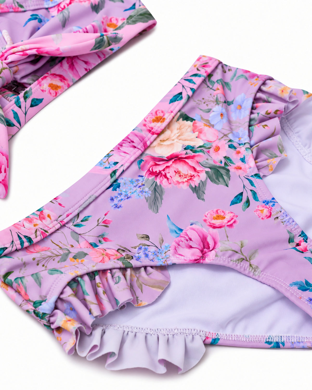 A close-up of a children's bikini bottom in light lavender with a colorful floral print, featuring ruffled trim along the leg openings, shown on a white background.