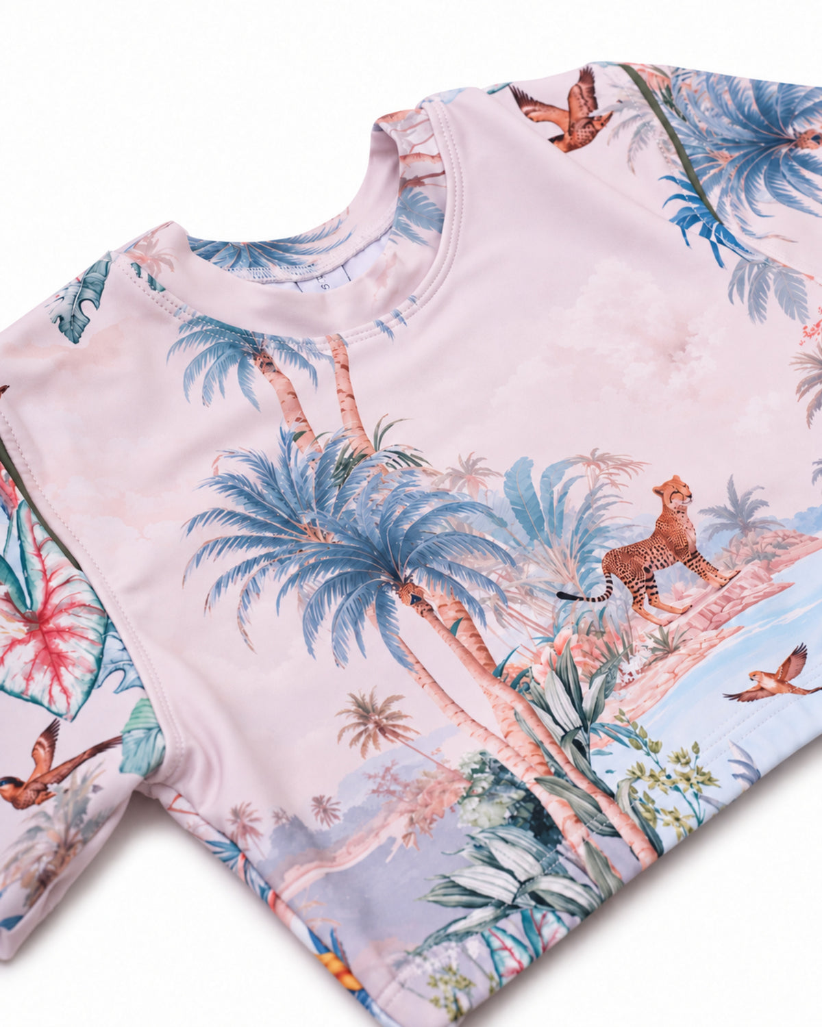 A close-up of a short-sleeve  crop rashie twith a light pink base and a colorful tropical print featuring blue palm trees, a cheetah, birds, and tropical foliage.