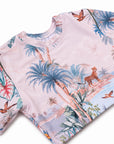 A close-up of a short-sleeve  crop rashie twith a light pink base and a colorful tropical print featuring blue palm trees, a cheetah, birds, and tropical foliage.